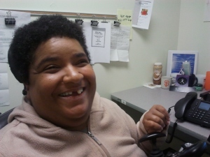 An African American woman with short black hair. She's inside an office and sitting nearby a table with a computer. She's wearing a beige pullover and smiling at the camera.